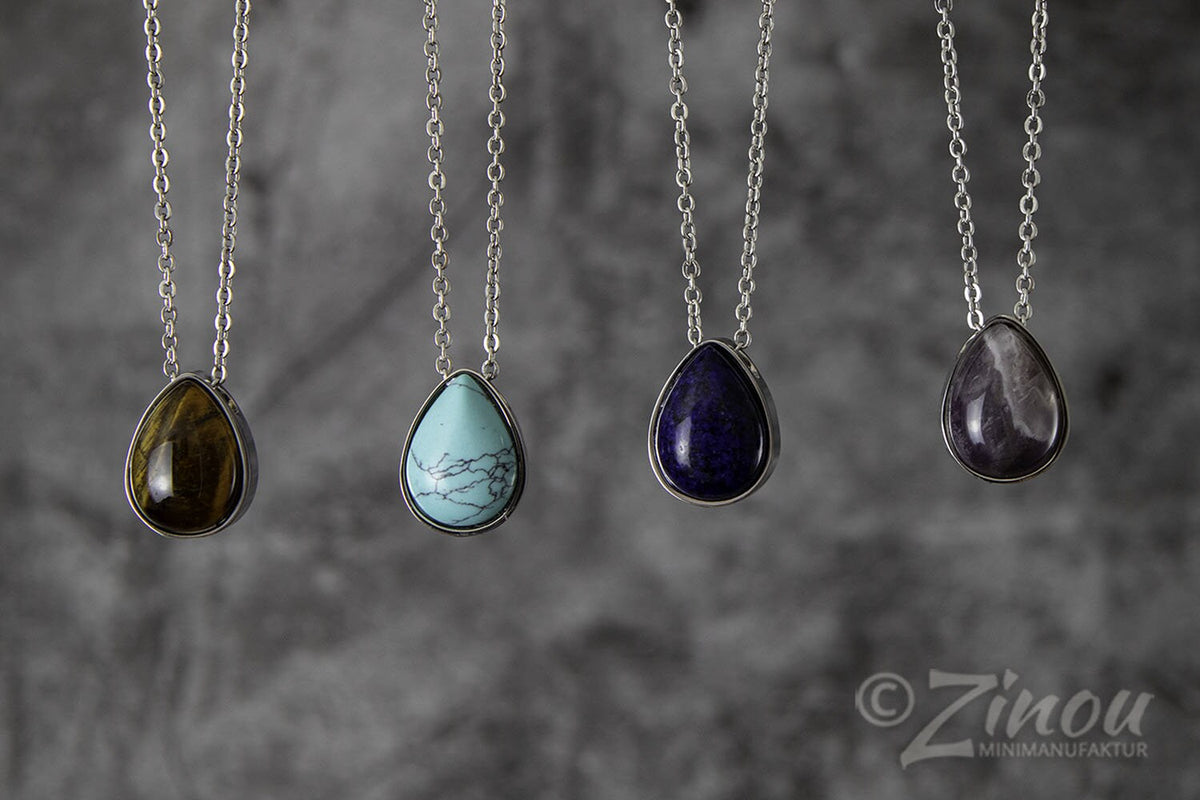 Vier silberne Tropfen-Urnenanhänger mit verschiedenen Halbedelsteinen – Tigerauge, Howlith, Sodalith und Amethyst – hängend an Silberketten vor grauem Hintergrund – Gedenkschmuck von Zinou Minimanufaktur