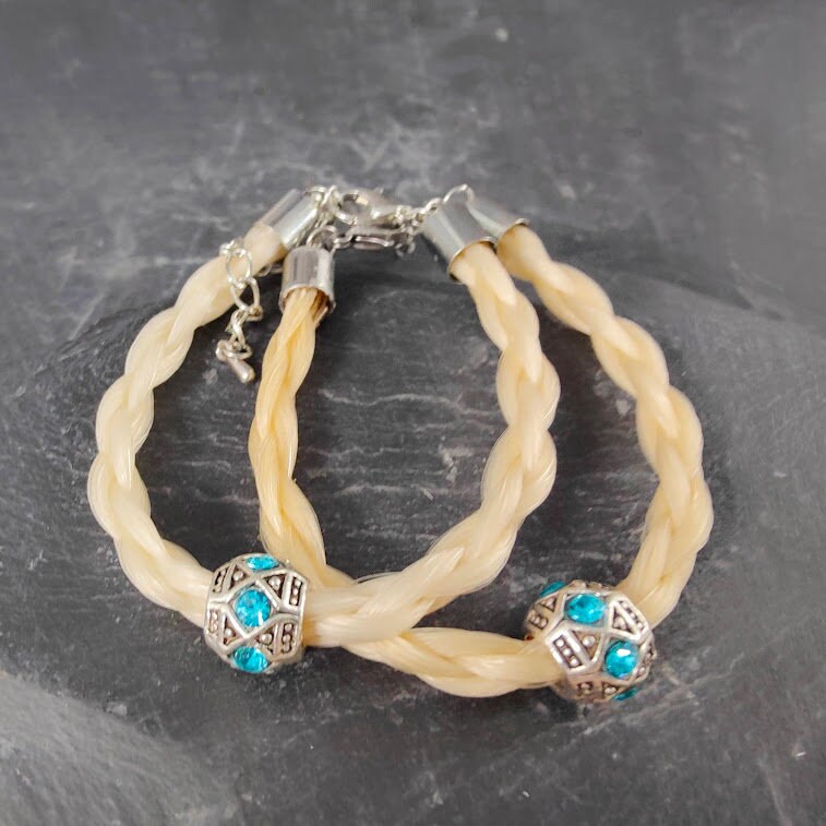 Zwei geflochtene blonde Pferdehaararmbänder mit silberner Perle und türkisafarbenen Strasssteinen auf Schieferstein, Seitenansicht mit Edelstahlverschluss – Pferdehaarschmuck von Zinou Minimanufaktur